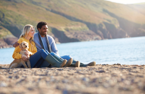 A couple with their dog sitting on a beach looking out at the water, and hills surrounding